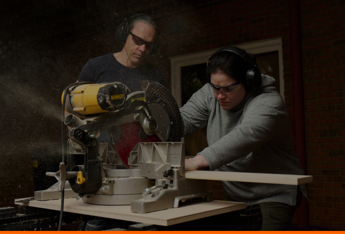 Caucasian male wearing PPE teaching Caucasian female wearing PPE how to use a chop saw to cut a piece of lumber