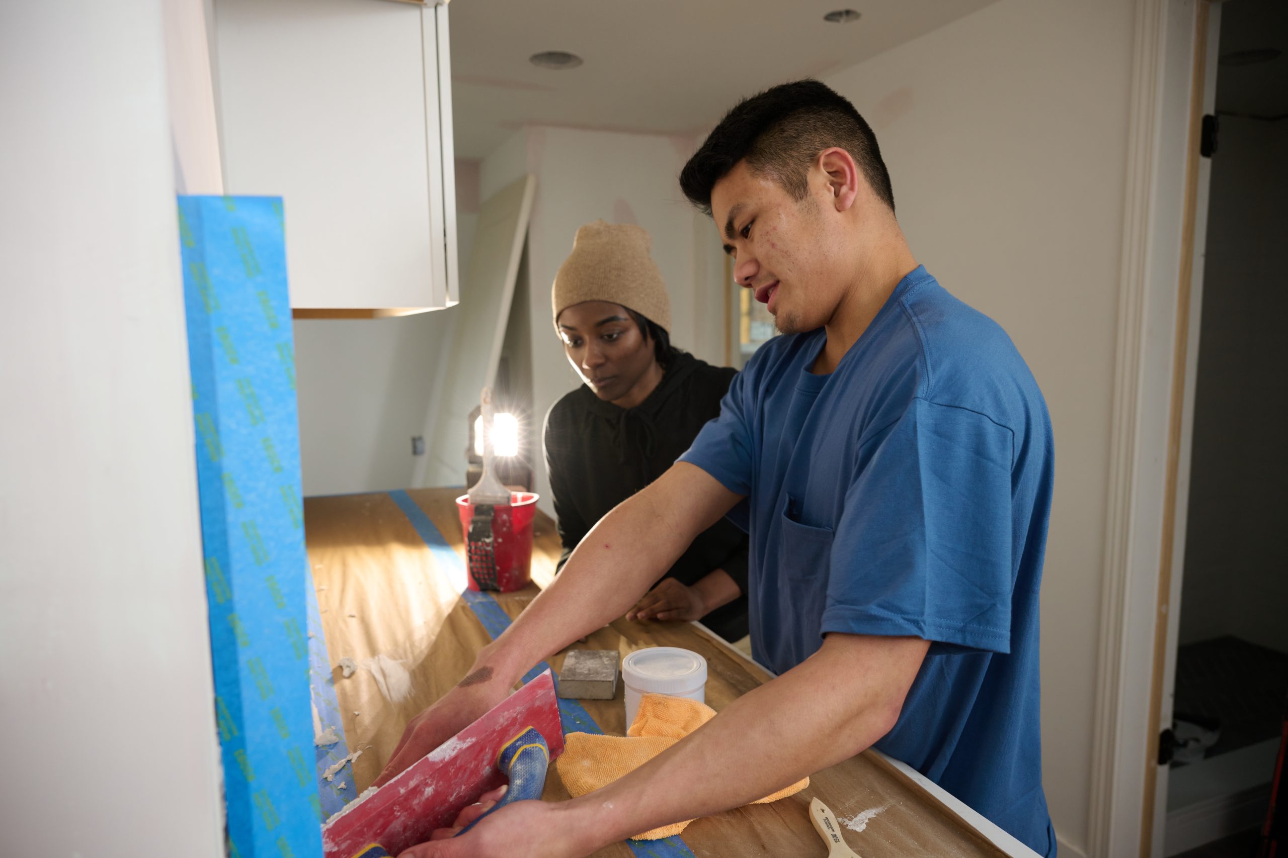Asian male teaching African American female how to apply grout to tile in a kitchen