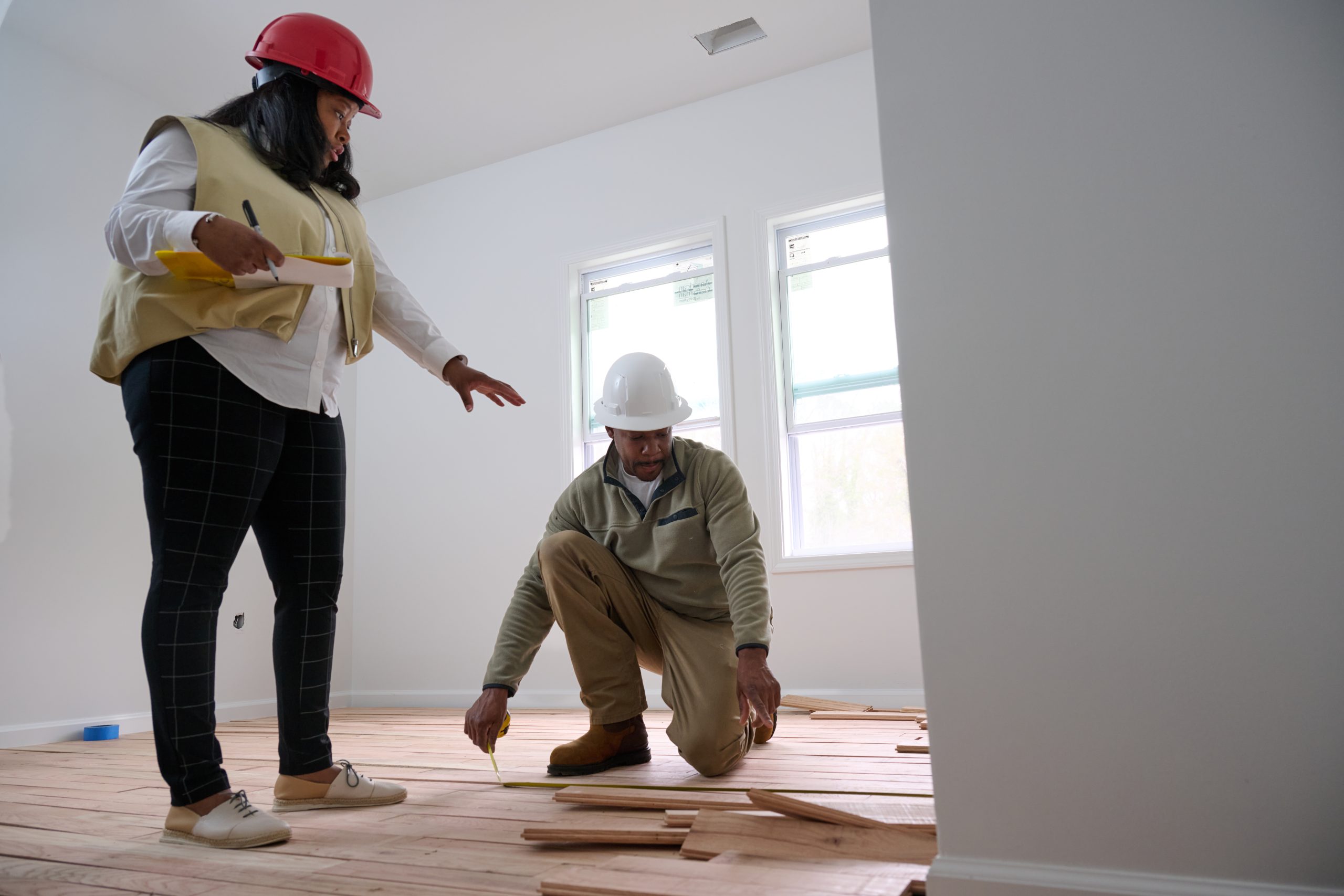 African American female in a hard hat discussing flooring with an African American male kneeling down in a hard hat