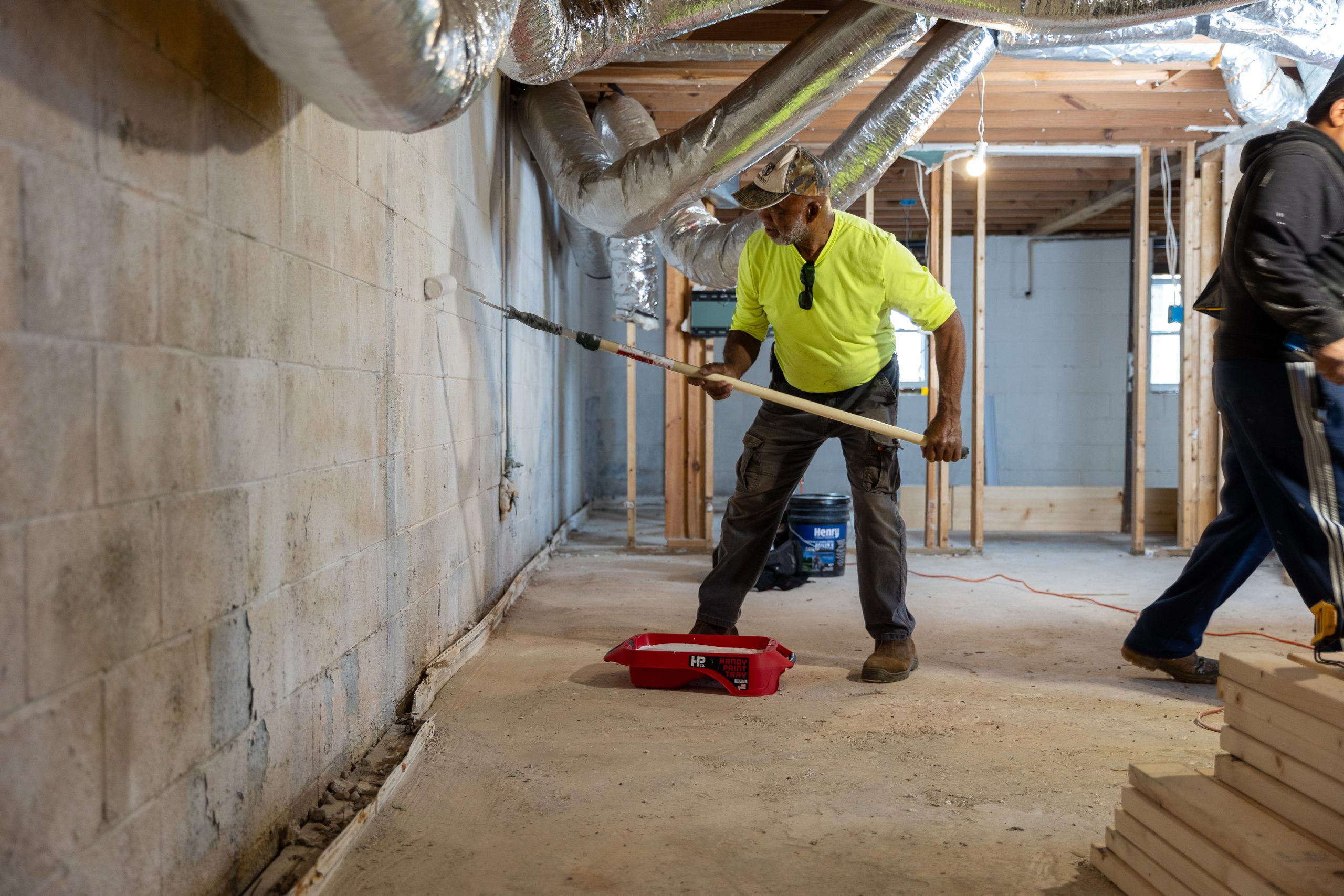African American male using a long paint roller to apply waterproofing barrier in a basement