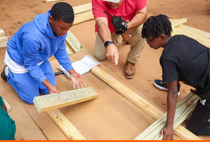 Two teens and an adult volunteer kneel outdoors, assembling a wooden frame from lumber boards during a community build project.